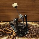 Černý basový pedál s kovovými mechanismy a řetězovým přenosem. Na horní části se nachází bílý plstěný tlumící prvek na kovovém rameni. Zařízení je um…