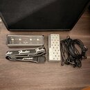 Na fotografii je kytarový pedál stříbrné barvy s nápisem Fender, vybavený několika ovládacími knoflíky a přepínači. Vedle něj je stříbrný pedál s per…