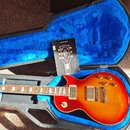 Elektrická kytara s trupem v červeno-žlutém sunburst provedení, umístěná v otevřeném kufru s modrým vnitřkem. Na hlavě kytary je bílý nápis Epiphone.…