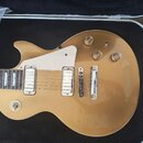 Zlatá elektrická kytara s tvarem single-cut, položená v černém tvrdém kufru. Nástroj má dva humbuckery s krémovými kryty, krémový pickguard a čtyři z…