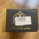 Černá krabice s nápisy a logy ve zlaté barvě. Na přední straně je nápis „lichtlaerm audio“ a geometrický symbol s písmenem „T“. Bílá nálepka obsahuje…