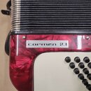 Detail akordeonu s červeným perleťovým povrchem a bílým panelem s černými knoflíky. Na červeném povrchu je stříbrný obdélníkový štitek s nápisem carm…