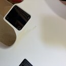 Detail zadní strany bílé elektrokytary s černým krytem elektroniky. Na krytu je vyražen nápis Squier. Povrch bílého laku vykazuje v okolí krytu vidit…