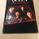 Fotografie znázorňuje černou sešitovou knihu s notami. Na obálce je bílý nápis QUEEN a pod ním červený text GREATEST HITS, v pravém horním rohu je dr…