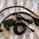 Sada černých kabelů a napájecí adaptér na světlém textilu s botanickým vzorem. Je zde vidět kabel s konektory 6,35 mm jack a MIDI, přičemž na jednom…