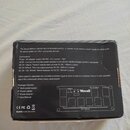 Fotografie zachycuje zadní stranu černého kartónového obalu od zdroje napájení. Na krabici je bílý text s popisem produktu, seznamem obsahu balení a…