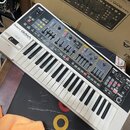 Bílo-šedý syntetizátor s klávesnicí a ovládacím panelem s mnoha posuvníky a tlačítky. Na horní části je nápis Roland GAIA SYNTHESIZER SH-01. Povrch p…