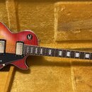 Elektrická kytara s tmelem v červeno-žlutém přechodu (sunburst) umístěná v hnědém plišovém pouzdře. Na hlavě je bílý nápis Epiphone a vložka s textem…