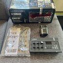 Na fotografii je elektronický modul Yamaha EAD10 v šedé barvě s ovládacími knofíky a displejem. Vedle něj je umístěn černý přívodní adaptér s nápisy…
