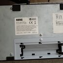 Fotografie zachycuje spodní stranu bílého elektronického zařízení s černými gumovými nožičkami. Na bílém štítku je uveden název značky KORG, model AX…
