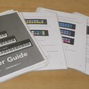 Fotografie zobrazuje několik listů papíru s textem a obrázky, které tvoří uživatelský manuál. Na předním listu je nápis User Guide, název modelu LAUN…