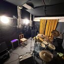 Fotografie zachycuje hudební studio s akustickými panely na stěnách a černým stropem. V prostoru je umístěna bicí souprava s černé barvou korpusů a z…