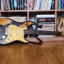 Elektrická kytara s černým povrchem, který vykazuje výrazné opotřebení a odřeniny odhalující světlý základ. Nástroj má žlutý pickguard, hnědý hmatník…