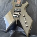 Elektrická kytara s špičatým korpusem v olivově zelené barvě a hnedým hmatníkem. Je vybavena dvěma černými humbuckery, z nichž na spodním je viditeln…