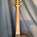 Zadní pohled na elektrickou kytaru z přírodního dřeva se světlým hnedem. Na zadní straně krku je kovový štít s nápisem Handcrafted in France. Hlavu k…