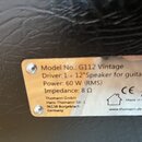 Detailní záběr na kovový štítek s technickými specifikacemi připevněný k černému povrchu z imitace kůže. Na štítku je text: Model No.: G112 Vintage,…