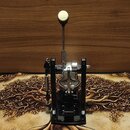 Černý basový pedál s kovovými mechanismy a bílým polstrováním na špičce beateru. Nástroj je umístěn na vzorované podložce s motivem stromu před dřevě…