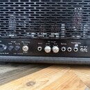 Detail zadní strany černého kytarového komba s mřížkou, za kterou jsou vidět elektronické lampy. Na ovládacím panelu jsou nápisy Power, FUSE T1 A TO…