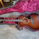 Akustická kytara s cutawayem v hnědém sunburst provedení, umístěná v černém tvrdém pouzdře s purpurovým polstrováním. Na hlavě hmatníku je nápis Tayl…
