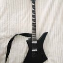 Černá elektrická kytara s ostře hranatým korpusem a černým popruhem. Na hlavě hmatníku je bílý nápis Jackson. Kytara má dva humbuckery, jeden ovládac…