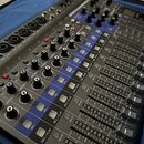 Šedý mixážní pult s modrými prvky a řadou posuvníků. Na panelu jsou viditelné nápisy jako GAIN, COMP, SEL, REC/PLAY a MUTE. Zařízení obsahuje početné…