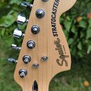 Fotografie zachycuje hlavu elektrické kytary z přírodního světlého dřeva s chromovanými ladičkami. Na povrchu je černý nápis Squier STRATOCASTER® by…