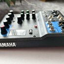 Modrý mixážní pult s černým plastovým rámem. Na zařízení jsou vidět nápisy YAMAHA, MG06 a označení vstupů jako PHONES, L a R. Povrch obsahuje několik…