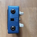 Detailní pohled na modrý kovový efekt pedál s kovovými konektory. Na šasi jsou nápisy INPUT, OUTPUT a 9V DC s označením polarity (+ -). Vlevo jsou vi…