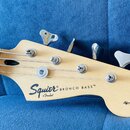 Detail hlavy světlé dřevěné basové kytary s nápisy Squier by Fender a BRONCO BASS™. Nástroj má čtyři chromované ladičky a čtyři struny. Povrch dřeva…