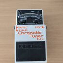Bílý kytarový ladič s černým spínacím pedálem, na kterém je vyražen nápis BOSS. Na horní části je analogový ukazatel s nápisy CHECK, BASS, GUITAR a C…