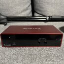 Červené audio rozhraní s kovovým povrchem. Na horní straně je nápis Focusrite a na přední hraně vlevo logo Focusrite. Jsou zde vidět dva výstupy s ná…