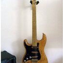 Elektrická kytara s přírodním dřevěným povrchem těla a hmatníkem. Má černý pickguard, tři snímače a stříbrné potenciometry. Na hlavě krku je viditeln…