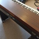 Digitální piano s klávesnicí a dřevěným povrchem v hnědé barvě. Na přední straně a nad klávesami je zlatý nápis YAMAHA. Nástroj stojí na černé kovové…