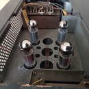 Fotografie zachycuje vnitřek elektronického zařízení, pravděpodobně zesilovače. V šasi jsou osazeny čtyři elektronky s kovovými vrछoly, z nichž dvě p…