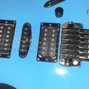 Detail modré elektrokitary s dvěma humbuckery a jedním singlovým snímačem. Na středovém snímači je svisle napsaný bílý nápis NFST. Kytara má černý mo…