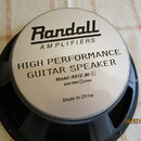 Kruhový kytarový reproduktor s černým rámem a stříbrnou středovou krytkou. Na krytce je černý nápis Randall AMPLIFIERS a text HIGH PERFORMANCE GUITAR…
