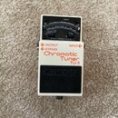 Bílý kytarový ladič s černým pedalem a displejem. Na zařízení jsou nápisy OUTPUT, BYPASS, INPUT a červeným písmem Chromatic Tuner TU-3. Na černém ped…