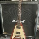 Elektrická kytara s červeným tělem ve tvaru V a krčkem z tmavého dřeva. Na bílém pickguardu jsou dva humbuckery a tři ovládací knofíky. Kytara je opř…