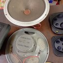 Fotografie zobrazuje několik reproduktorových jednotek položených na hnědém stole. Horní reproduktor má šedou membránu a bílý kovový rám. Spodní repr…