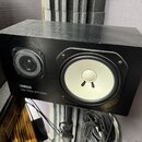 Černý studiový monitor s bílou membránou basového reproduktoru a černým výškáčem za mřížkou. Na přední straně je bílý nápis YAMAHA a pod ním model NS…