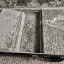 Otevřený kufřík s vnitřním polstrováním z šedého plyšového materiálu. V interiéru jsou vidět přihrádky pro ukládání příslušenství. Vnější strana kufř…