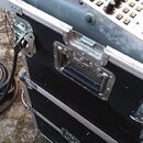 Detailní pohled na část kontrolního panelu s tlačítky a potenciometry, který je umístěn v černém transportním kufru s hliníkovými hranami a madlem. P…