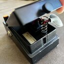 Černý kytarový pedál s otevřeným horním krytem, který odhaluje vnitřní mechanismus s pružinou a bílým polstrováním. Uvnitř je vidět bílá nálepka s ná…