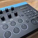 Černý elektronický bicí modul s řadou ovládacích prvků a displejem. Na horním okraji je modrý štítek s nápisem Roland TR-8S. Zařízení obsahuje několi…