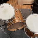 Tři bubny s povrchem z brownish-orange dřeva s výraznou strukturou zrn. Všechny tři mají bílé blány s černým logem REMO. Bubny jsou osazeny chromovan…