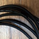 Černý audio kabel s matným povrchem, který je složen do několika oblouků na dřevěném podkladu. Na kabelu je bílý nápis the sssnake www.thomann.de a z…