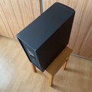 Černý hranatý reproduktor umístěný na dřevěné stoličce. Na zadní straně je vidět červený vypínač a část žlutého štítku. Povrch je matný s viditelným…