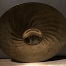 Zlatavý mosazný činel s výrazným kovánívým vzorem v podobě spirálovitých řad důlků. Povrch vykazuje známky opotřebení, oxidaci a drobné nečistoty. Na…