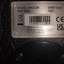 Detailní záběr na stříbrný typový štítek připevněný k černému texturovanému povrchu. Na štítku je uveden nápis MODEL: MX112R, 80W RMS, LEAD 1x12" a 1…