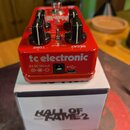 Červený efekt pedál značky tc electronic umístěný na bílé krabici s nápisem HALL OF FAME 2 REVERB. Pedál má dva černé ovládací knoflíky a nápisy TONE…